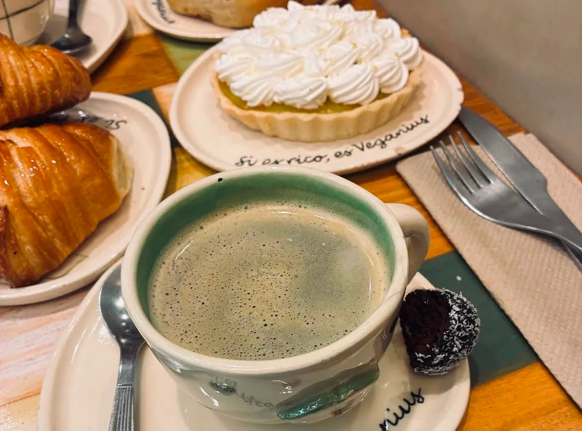 Café de especialidad en taza verde con tarta de limón y merengue de fondo; el plato dice “Si es rico, es Veganius”